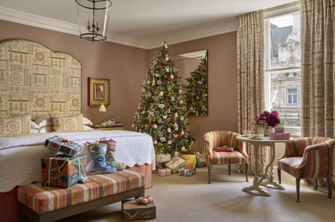 Deluxe Junior Suite 303 decorated for Christmas at Haymarket Hotel. Presents and stockings sit at the end of the bed. In the corner there is a decorated Christmas tree with presents underneath. In front of the window, there is a table with presents and Champagne and two chairs either side