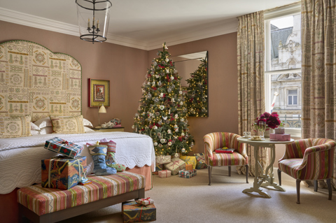 Deluxe Junior Suite 303 decorated for Christmas at Haymarket Hotel. Presents and stockings sit at the end of the bed. In the corner there is a decorated Christmas tree with presents underneath. In front of the window, there is a table with presents and Champagne and two chairs either side