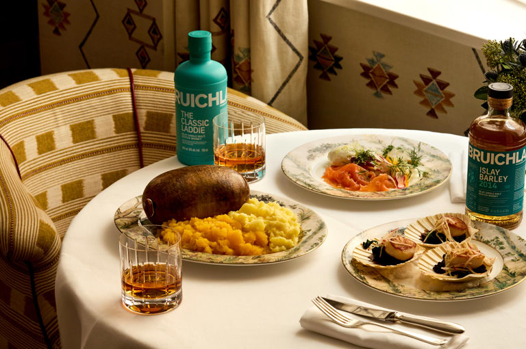 A table set for Burns Night dinner with Bruichladdich at Covent Garden Hotel. There is a plate of haggis being cut open, plates of scallops and smoked salmon and glasses of whisky and Bruichladdich bottles.