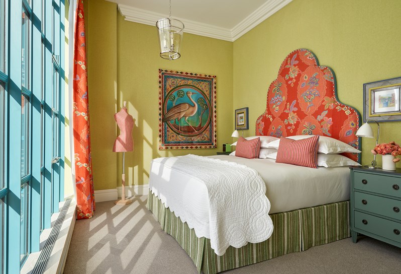 A lime green walled room with floor ceiling windows opposite a bed with a headboard upholstered in orange 'Chainstitch' fabric. There are two pink and orange striped cushions on the bed which match a mannequin in the left hand corner. There is a painting of a large bird on the wall to the left of the bed and a turquoise bedside table to the right.