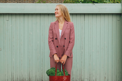 A portrait shot of food writer, journalist and broadcaster Alexandra Dudley, a young woman with long blonde hair. She is dressed in a red gingham suit, holding a red leather bag filled with kale