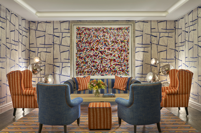 An image of the seating area in the Sandra Blow private events room at The Soho Hotel. The image shows a sofa in a blue and orange colour scheme under a piece of art, with two orange armchairs flanking the sofa and two blue arm chairs at the front facing away from the camera. 