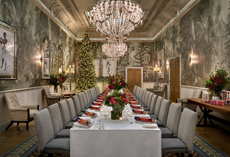 A long table setup vertically in the Shooting Gallery seating around 15 guests on each side. There are Christmas crackers on each plate alongside flowers and candles lit up in the centre. There is a Christmas tree in the corner.