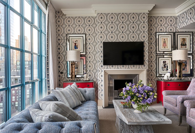 Two blue and red patterned armchairs in front of a coffee table opposite a blue sofa. On the back of the room, a fireplace and TV on the wall, a red chest of drawers and dog lamp on either side.
