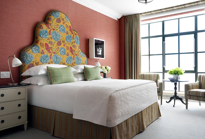 A double bed with a yellow, blue and red patterned headboard and two green cushions. On the right there are two striped armchairs in front of floor-to-ceiling windows.