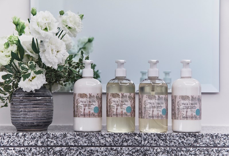 Four bottles of Tall Trees bath & body products lined up next to a vase of white flowers on marble surface.