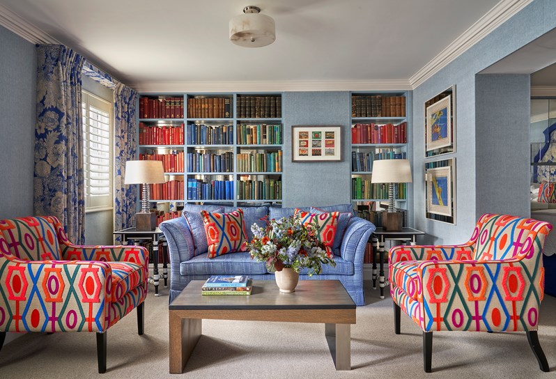 Seating area shot of room 15, the Marylebone Room at Dorset Square Hotel. The image shows a blue sofa in the centre in front of a colourful bookshelf wall full of books. In front of the sofa, there is a coffee table with patterned armchairs on either side.
