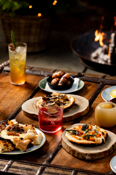 A selection of fire-cooked dishes and cocktails sit on a wooden coffee table