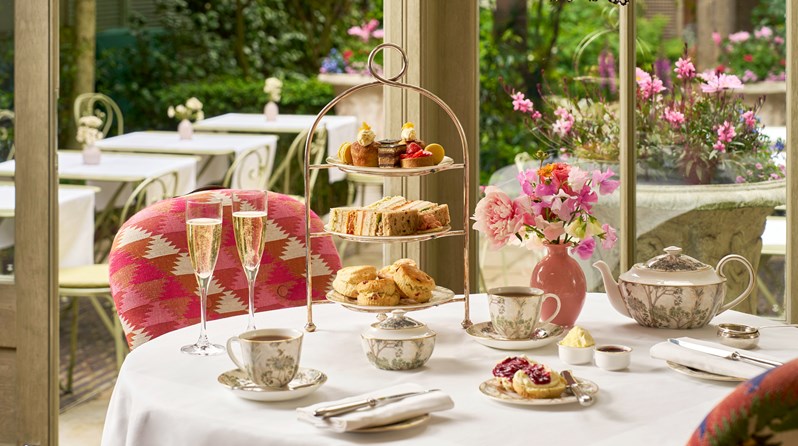 Afternoon tea set up on a table for two in The Orangery. A cake stand, with mini cakes, finger sandwiches and scones. Two cups of tea, a sugar bowl and teapot and a plate of jams and creams. A plate with a scone on.