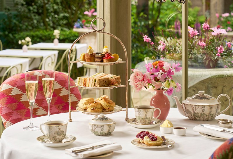 Afternoon tea set up on a table for two in The Orangery. A cake stand, with mini cakes, finger sandwiches and scones. Two cups of tea, a sugar bowl and teapot and a plate of jams and creams. A plate with a scone on.
