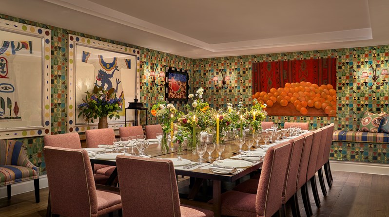 The newly photography Vanessa Bell Room setup for  dinner with orange chairs. There is beautiful bright artwork on the walls and flowers decorating the table with glasses, plates, cutlery and nakpins
