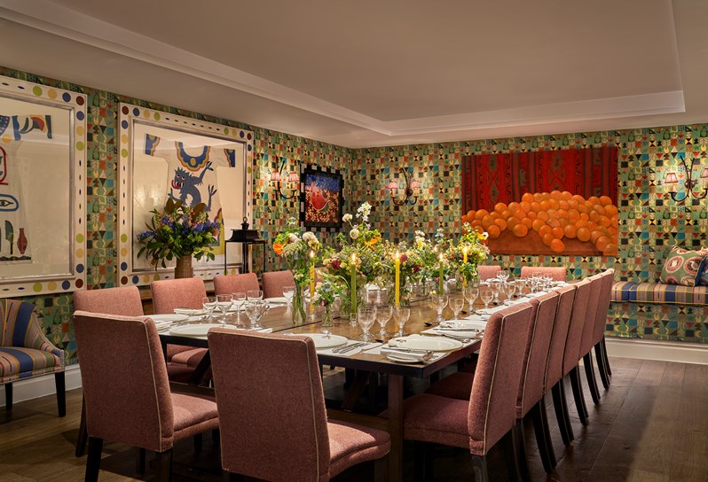 The newly photography Vanessa Bell Room setup for  dinner with orange chairs. There is beautiful bright artwork on the walls and flowers decorating the table with glasses, plates, cutlery and nakpins