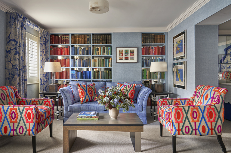 Seating area shot of room 15, the Marylebone Room at Dorset Square Hotel. The image shows a blue sofa in the centre in front of a colourful bookshelf wall full of books. In front of the sofa, there is a coffee table with patterned armchairs on either side.