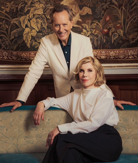 Christine Baranksi dressed in a blouse and black trousers sits on a blue chez long. Richard E. Grant stands behind her dressed in a cream suit