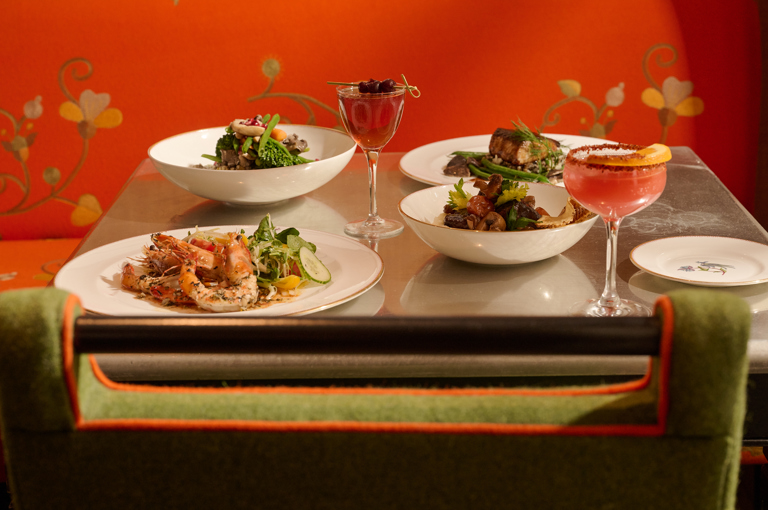 Close up shot of a table of main courses including, prawns, two salads and a beef fillet with two cocktails on a table at The Crosby Bar