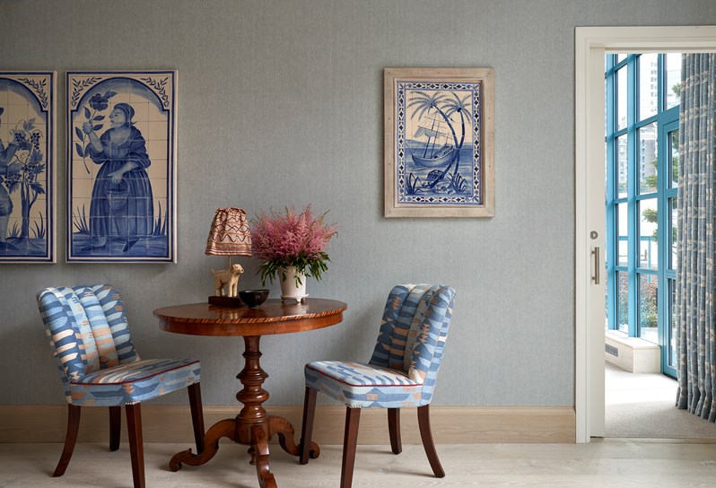 Two blue patterned chairs with a wooden table in the middle with a doggy lamp and flowers on. On the blue wall behind, there are three blue and white tile mosaics and a door to the next room on the right.