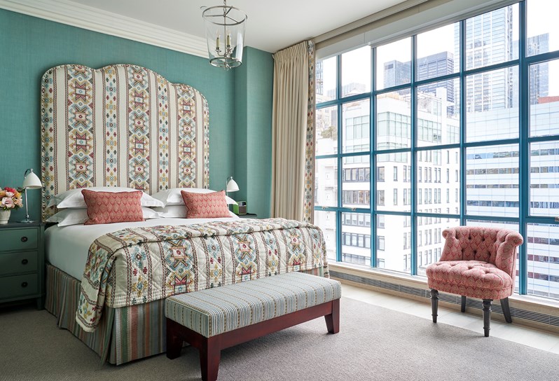 A bedroom with a geometric statement headboard that matches a throw at the end of the bed and two pink and white cushions that match a tub chair in front of floor to ceiling windows. Two blue bedside tables either side.