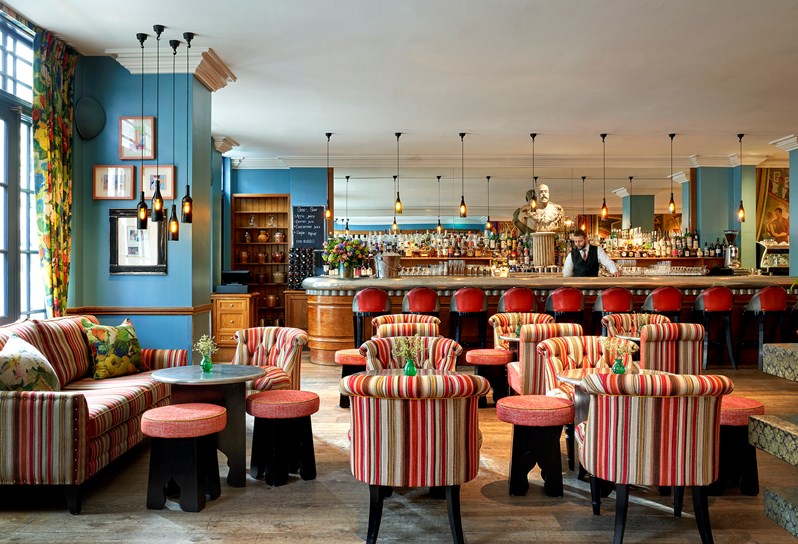 Oscar Bar - there are red striped sofas and armchairs with small bar tables. Behind is a long pewter bar with red leather bar seats. A barman is standing behind.
