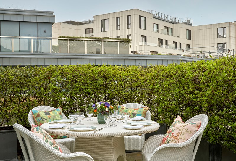 A white wicker garden table sits on an outdoor terrace and is laid for lunch with plates and glasses. Patterned cushions sit on each of the four chairs