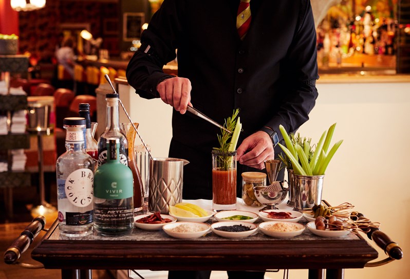 A 'Bloody Mary' trolley. There are several small bowls of garnishes, bottles of different spirits, a jug of tomato juice and pots of rosemary and celery on the trolley. Behind, the bottom half of a bartender can be seen placing a celery stick into a Bloody Mary.