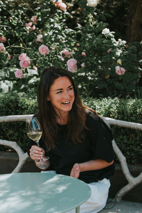 A portrait shot of Emily Harman (VinaLupa) sat on the roof terrace at Ham Yard Hotel, holding a glass of white wine against a floral backdrop.