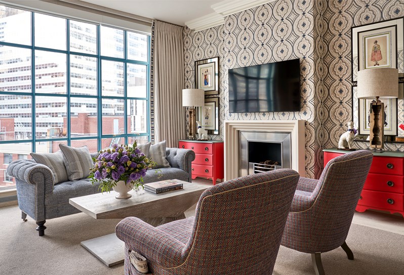Two blue and red patterned armchairs in front of a coffee table opposite a blue sofa.  A fireplace and TV on the wall in the middle and two framed drawings hung above a red chest of drawers and dog lamp on either side.
