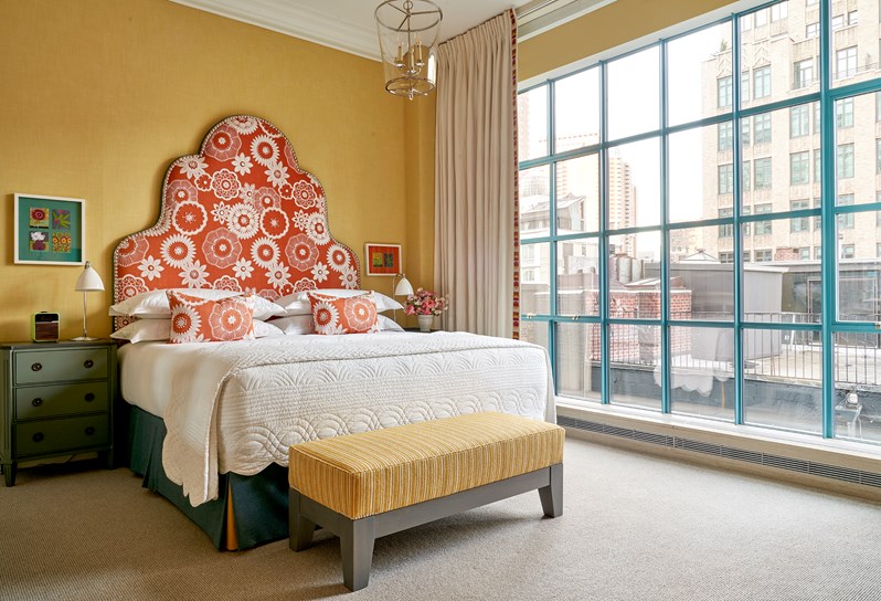 A bed with an orange headboard and cushions with white floral stitching in front of a yellow wall with a yellow ottoman at the end. Floor to ceiling windows on the right hand side.