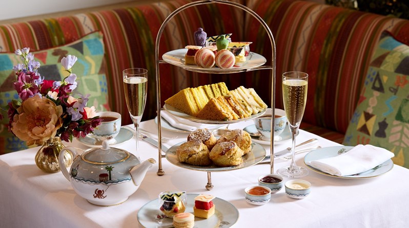 A table displaying Kit Kemp Design Thread Afternoon Tea at The Whitby Bar. There is a three-tiered stand holding a layer of scones, sandwiches and on top, a selection of miniature pastries inspired by Kit Kemp's designs. There are two glasses of Champagne, teapot and teacups with a small plate of the mini pastries also on the table.
