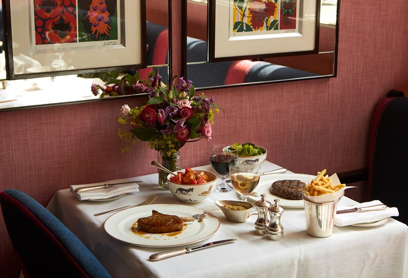 Brumus table set up for two seen from a diagonal angle with a pork chop and a side of tomato salad on the left side and a steak with a side of green leaf salad and french fries with two glasses of wine red and white on the the right side.