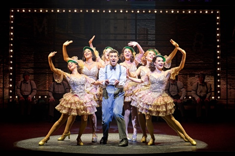 A smartly dressed man stands singing in the middle of a group of women dancing around him on stage in The Producers