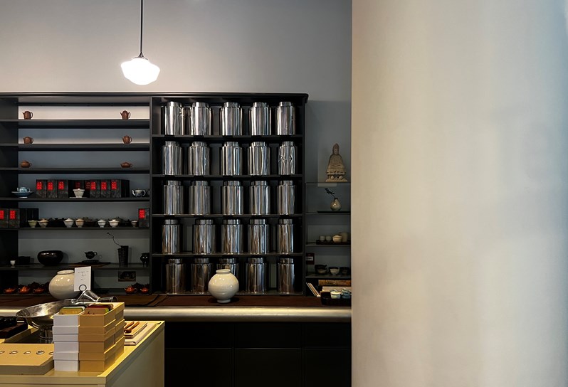 Inside Yushu Tea Gallery. There is shelving unit along the back wall housing metal containers, tea pots and tea cups. There is a work top in front