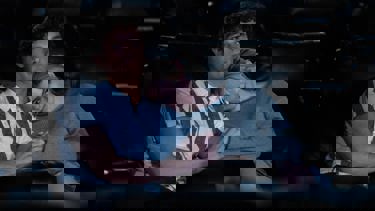 Still from the film 'Regretting You'. A teenage couple sitting on cinema seats. The girl leans against a the boy, with her head on his shoulder as they hold hands