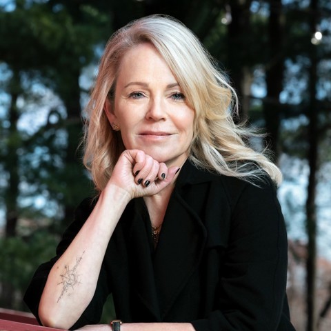 A headshot of author and podcaster Dani Shapiro. She is stood outside, resting her chin on her arm leaning on a wooden beam. Trees can be seen out of focus behind her