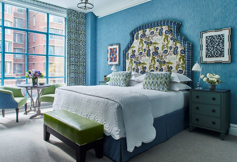 A blue and green bedroom with a large statement headboard a footstool upholstered in bright green leather and a round table with two tub chairs