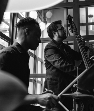 A black and white landscape image of a band playing jazz music, in a centre stands a guy with a upright bass and on the left a man playing the drums in the Warren Street Bar & Restaurant at Warren Street Hotel.