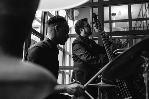 A black and white landscape image of a band playing jazz music, in a centre stands a guy with a upright bass and on the left a man playing the drums in the Warren Street Bar & Restaurant at Warren Street Hotel.