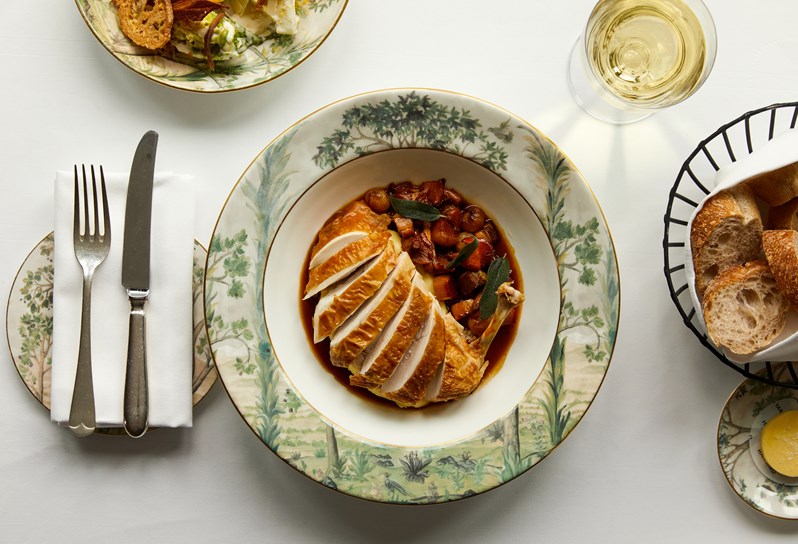 An image of a table with main course chicken dish in a bowl, at Brasserie Max restaurant at Covent Garden Hotel. The table also has a glass of wine and bread basket with butter. This image is to promote dining collaboration with The Producers theatre production.