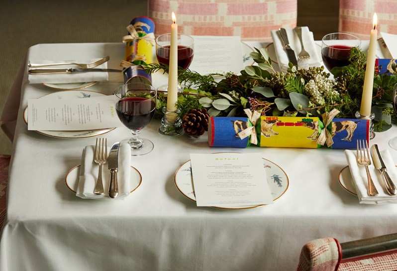 A closeup of a table setup for festive dining. There are Christmas crackers, candles, and plates with menu's being laid out, alongside a glass of red wine each. The crockery being used is Mythical creatures.