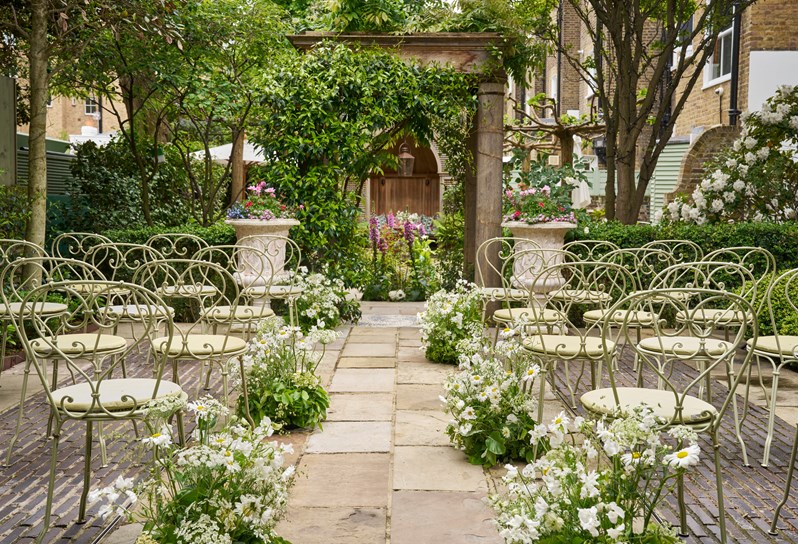 A wedding ceremony set up in a garden. There are rows of green metal chairs with white flowers growing from either side of the aisle. There is a stone archway with greenery growing over it.