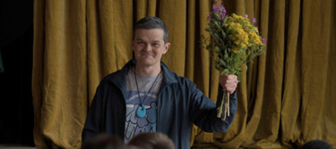 Still from the film 'I Swear'. A man stands smiling holding a bunch of flowers up in front of a yellow curtain.