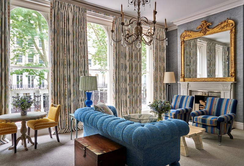 Seating area and fireplace shot of room 101, the Knightsbridge Suite at Knightsbridge Hotel. The image shows a diagonal shot from the bed area across the seating area, showing the blue sofa in the middle of the room facing towards two blue striped armchairs with a low coffee table in the center. A fireplace sits behind the armchair.