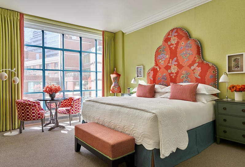 A bed with a headboard upholstered in orange Chainstitch fabric, matching the mannequin next to it, and two red striped cushions and an orange ottoman at the end. Two pink checked tub chairs and a coffee table in front the windows.