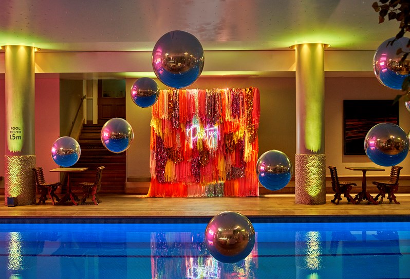 10 large Metallic looking baubles hanging from the ceiling over the pool with a neon-lit party board behind. There are two poolside tables visible and a sign saying the pool depth of 1.5 meters.