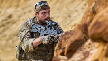 A still from the film 'Fuze'. A man dressed in camouflage army uniform crouches behind rocks in a desert setting, holding a large gun