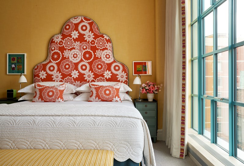 A bed with an orange headboard and cushions with white floral stitching in front of a yellow wall with a yellow ottoman at the end. Floor to ceiling windows on the right hand side.