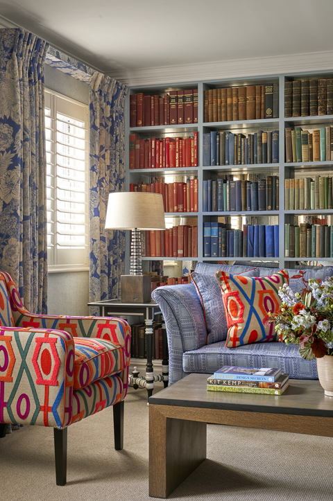 Portrait image of the seating area shot of room 15, the Marylebone Room at Dorset Square Hotel. The image shows a blue sofa in the centre in front of a colourful bookshelf wall full of books. In front of the sofa, there is a coffee table with patterned armchairs on the left side.