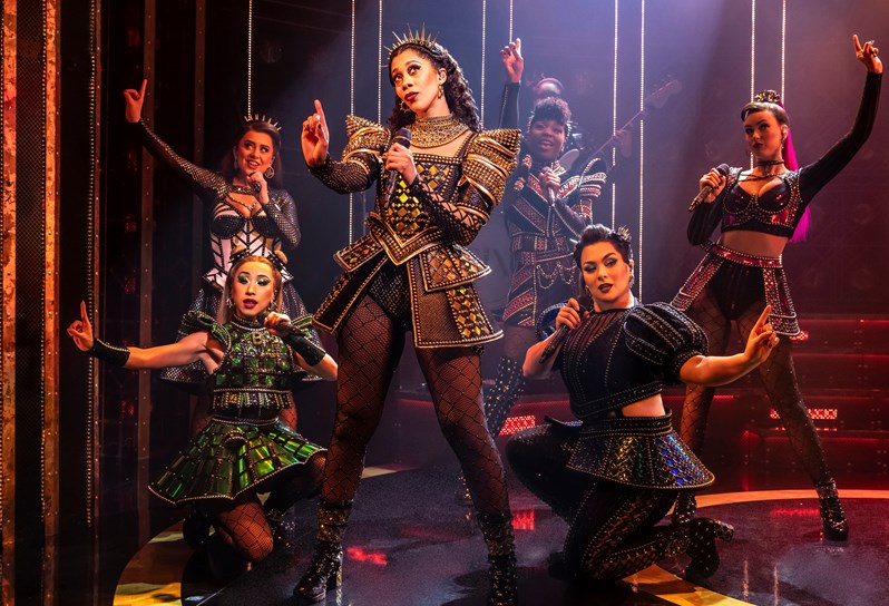 The six lead cast members of SIX the Musical. One woman stands in the centre, pointing her finger and holding a microphone. Five other cast members crouch and stand around her singing into microphones. All are dressed in shiny, structured dresses and wearing crowns.