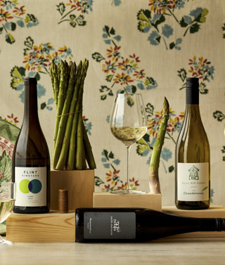 Wide shot of asparagus with wine bottles, glasses and a hand-painted ceramic plate set against and floral fabric backdrop