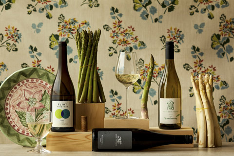 Wide shot of asparagus with wine bottles, glasses and a hand-painted ceramic plate set against and floral fabric backdrop