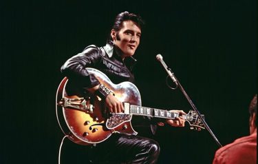 A still from the film "Epic Elvis Presley in Concert" showing Elvis Presley sitting in front of a microphone with a guitar.